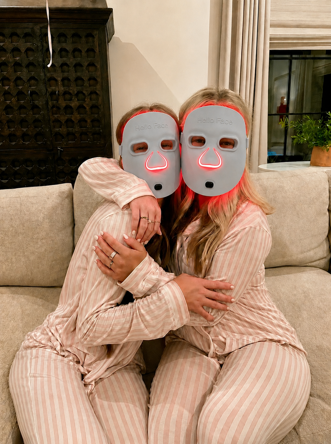 Two sisters in matching pink-striped pajamas wearing Louvaine LED masks together on the sofa
